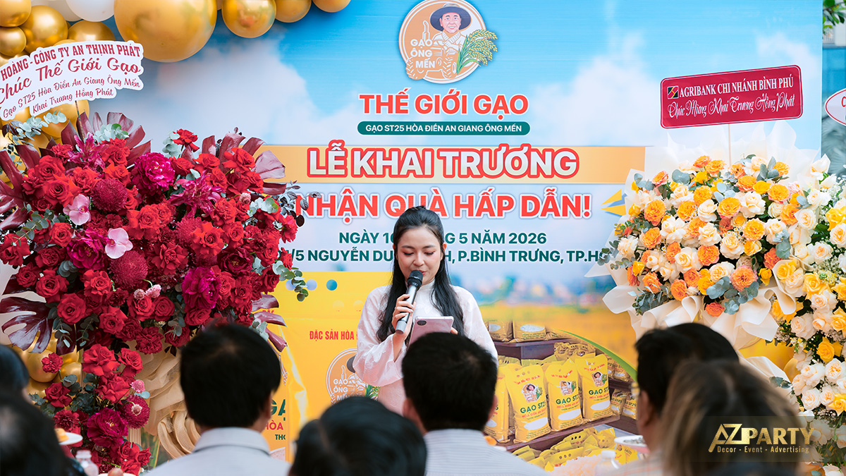 MC dẫn chương trình lễ khai trương Thế Giới Gạo mặc áo dài trắng cầm mic trước backdrop và hoa chúc mừng