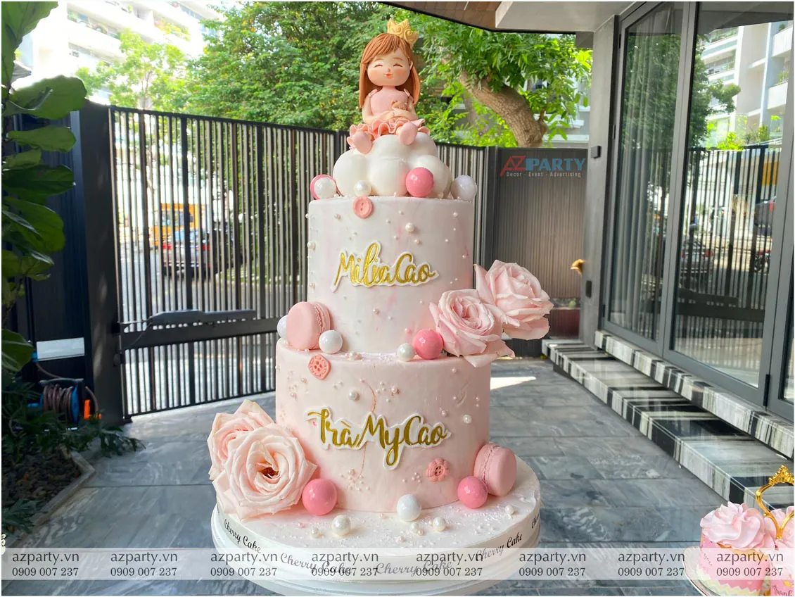 Cận cảnh chiếc bánh kem sinh nhật 2 tầng màu hồng nhạt xinh xắn trang trí hoa tươi, macarons và búp bê công chúa trên đám mây của bé Milia Cao và Trà My Cao.