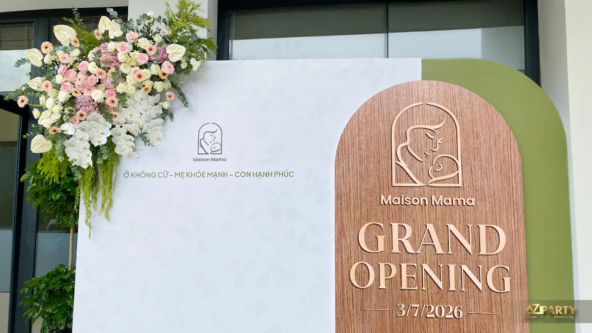 Cận cảnh backdrop Grand Opening Maison Mama với bảng gỗ bo vòm, logo thương hiệu và hoa tươi tone trắng hồng