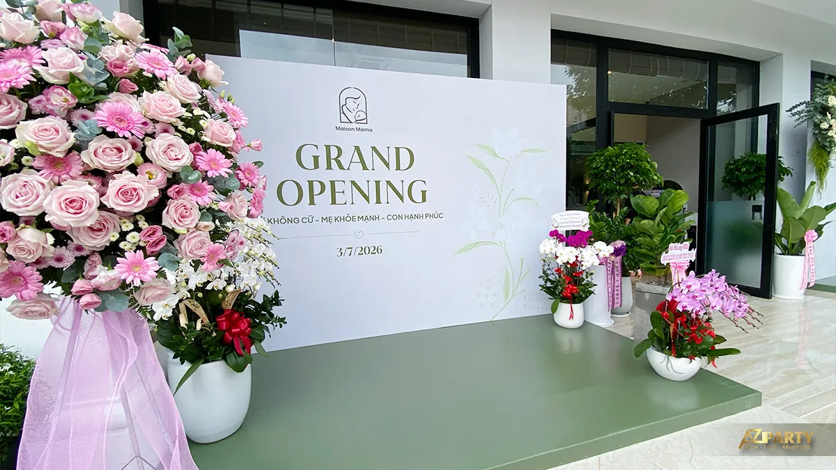 Backdrop bên trái lễ khai trương Maison Mama với chữ Grand Opening, sàn xanh và hoa chúc mừng tại Quận 7
