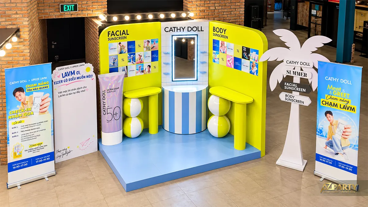 Toàn bộ khu check-in Cathy Doll từ góc cao với sàn xanh, hai bàn trưng bày và standee sự kiện