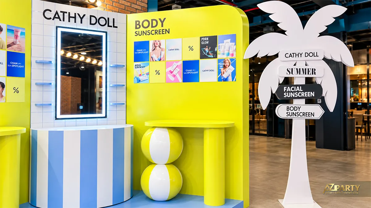Vách kem chống nắng cơ thể và biển chỉ dẫn hình cây dừa màu trắng cạnh booth Cathy Doll