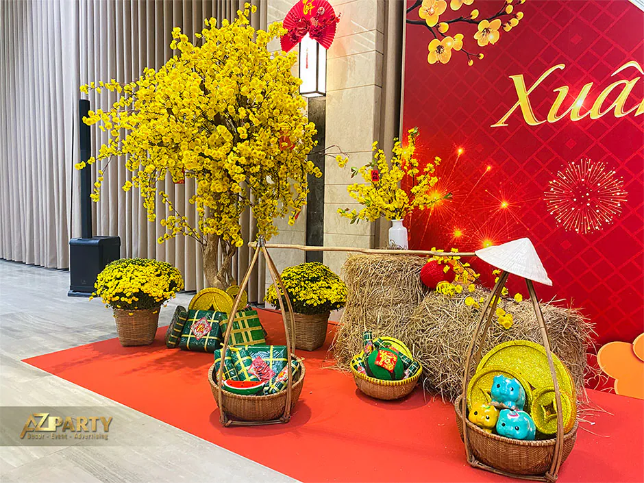 Chi tiết trang trí tiểu cảnh Tết gồm cây mai vàng lớn, giỏ tre đựng mô hình bánh chưng dưa hấu, đống rơm và chiếc nón lá mộc mạc.
