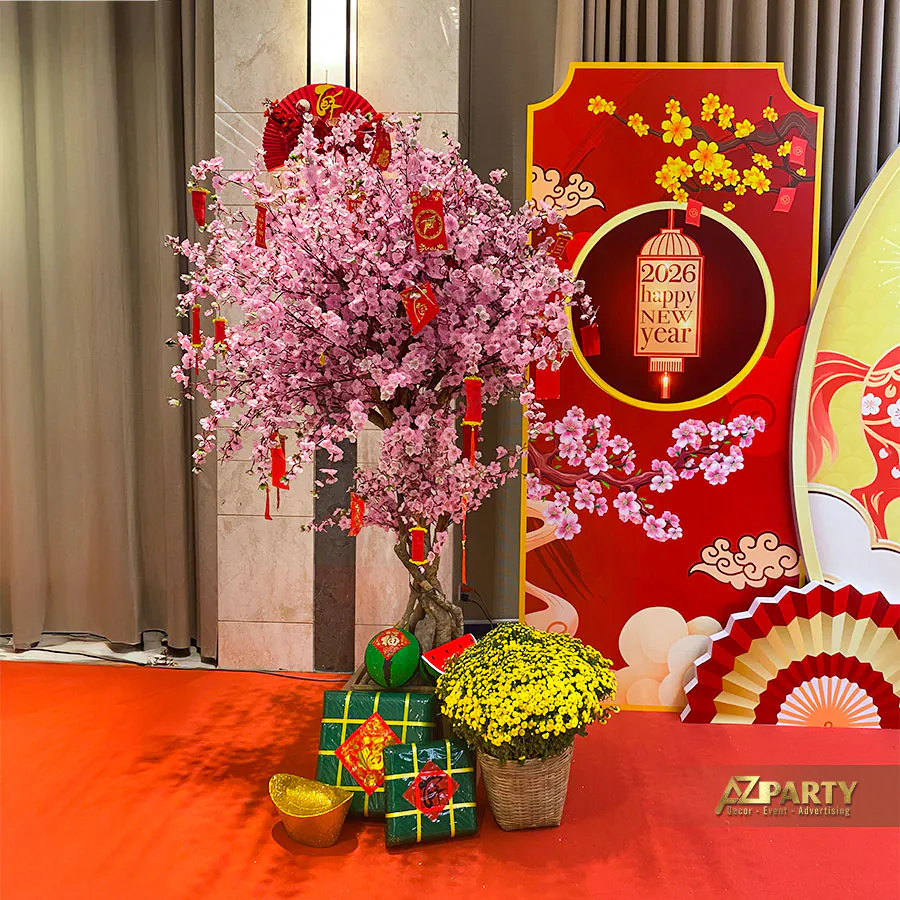 Cây hoa đào hồng đào rực rỡ treo phong bao lì xì đỏ may mắn, dưới gốc bày các mô hình bánh chưng, thỏi vàng và chậu cúc mâm xôi vàng óng.
