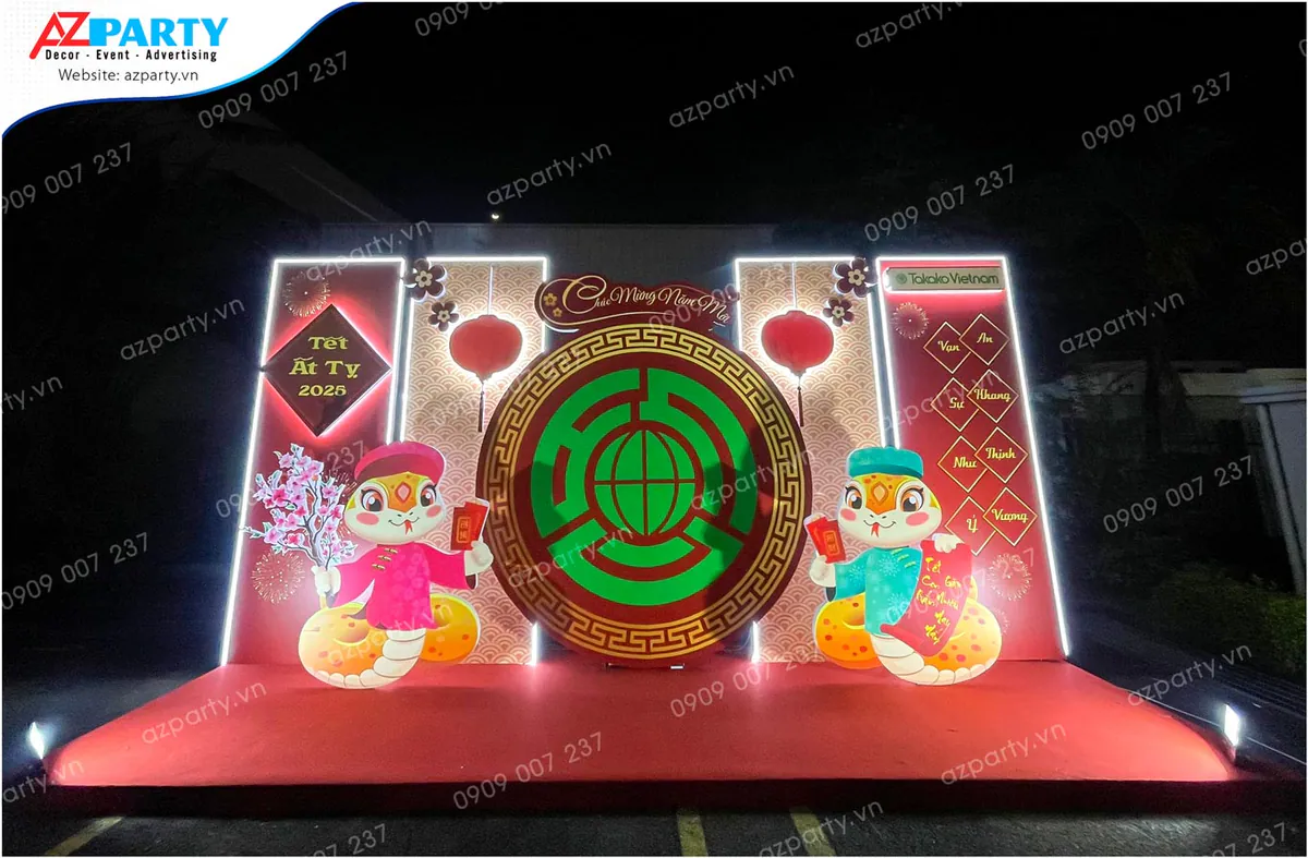 Trang trí tiểu cảnh Tết Ất Tỵ 2025 tại công ty Takako Vietnam với backdrop đỏ rực rỡ, hệ thống đèn LED sáng nổi bật, hai mô hình linh vật rắn ngộ nghĩnh cùng các câu chúc Tết truyền thống