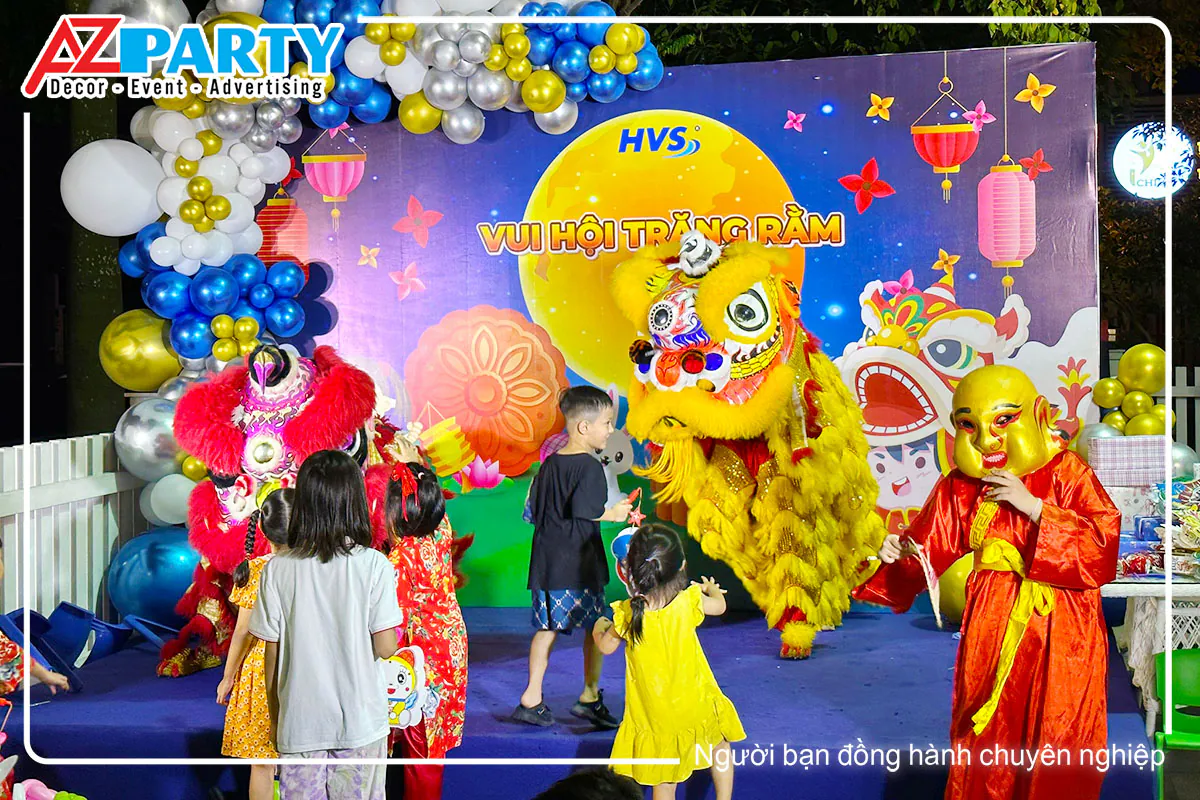 Đội múa lân đỏ và vàng biểu diễn trước sân khấu, các em nhỏ và ông Địa mặt nạ vàng xem múa lân tại Trung thu HVS