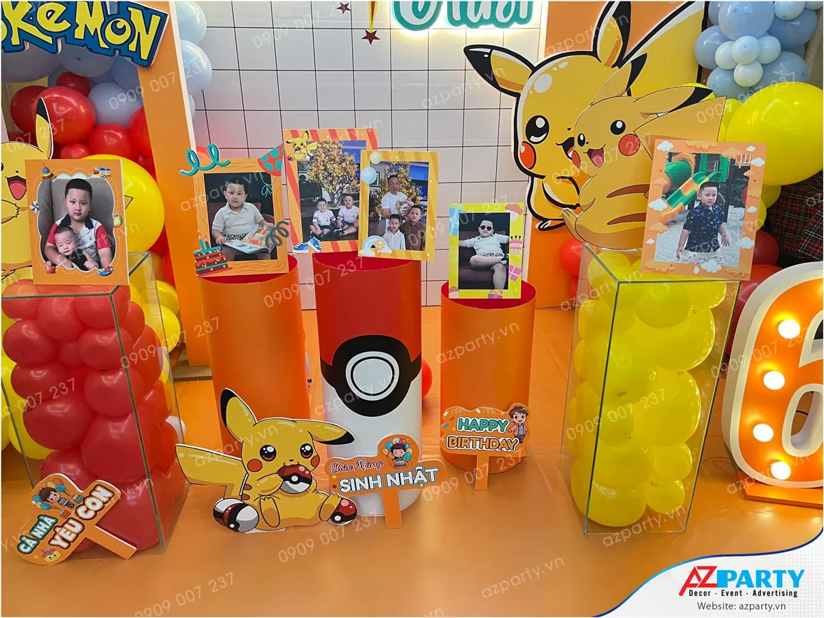Chi tiết bàn gallery sinh nhật bé Jonathan với các đôn trụ tròn in hình quả cầu Pokeball làm bệ đỡ khung ảnh, hộp mica chứa bóng bay vàng đỏ và nhiều hình cắt Pikachu ngộ nghĩnh.