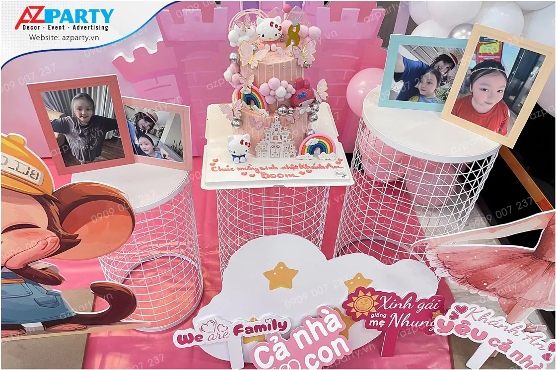 Chi tiết bàn gallery tiệc sinh nhật bé Khánh An với chiếc bánh kem Hello Kitty màu hồng dễ thương, các khung ảnh kỷ niệm gia đình và các biển hashtag ngộ nghĩnh.