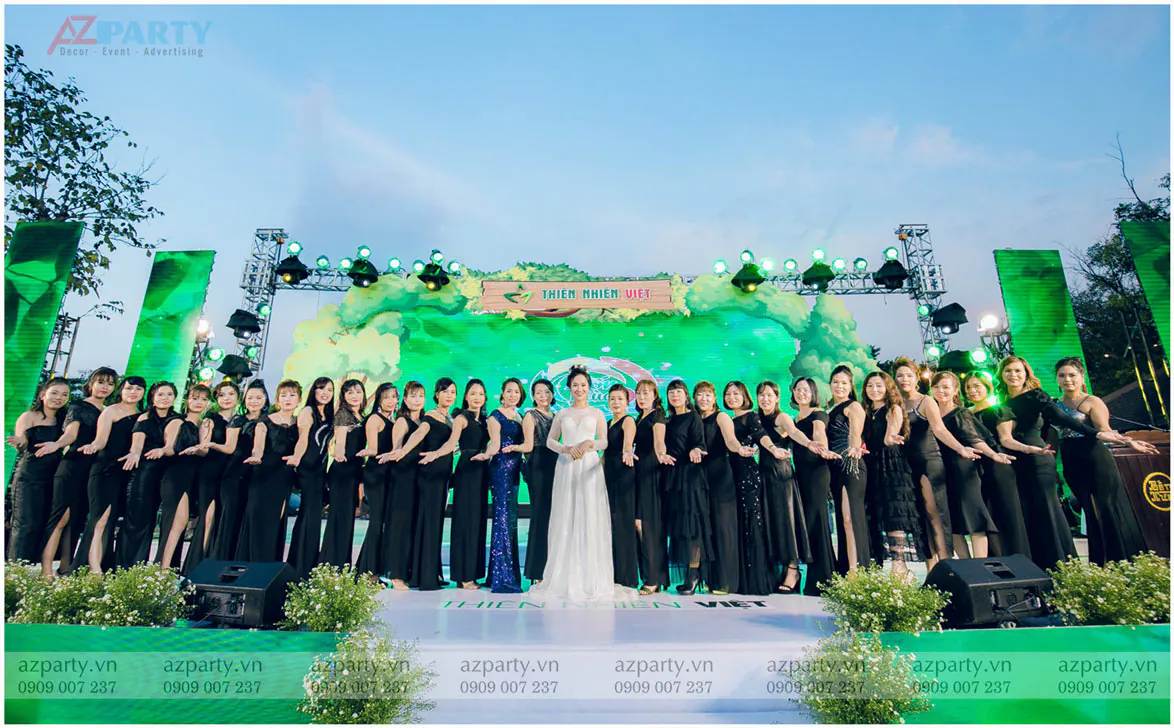 Tập thể đại lý Thiên Nhiên Việt mặc đầm dạ hội đen đồng điệu chụp ảnh cùng người sáng lập mặc đầm trắng trên sân khấu ngoài trời.