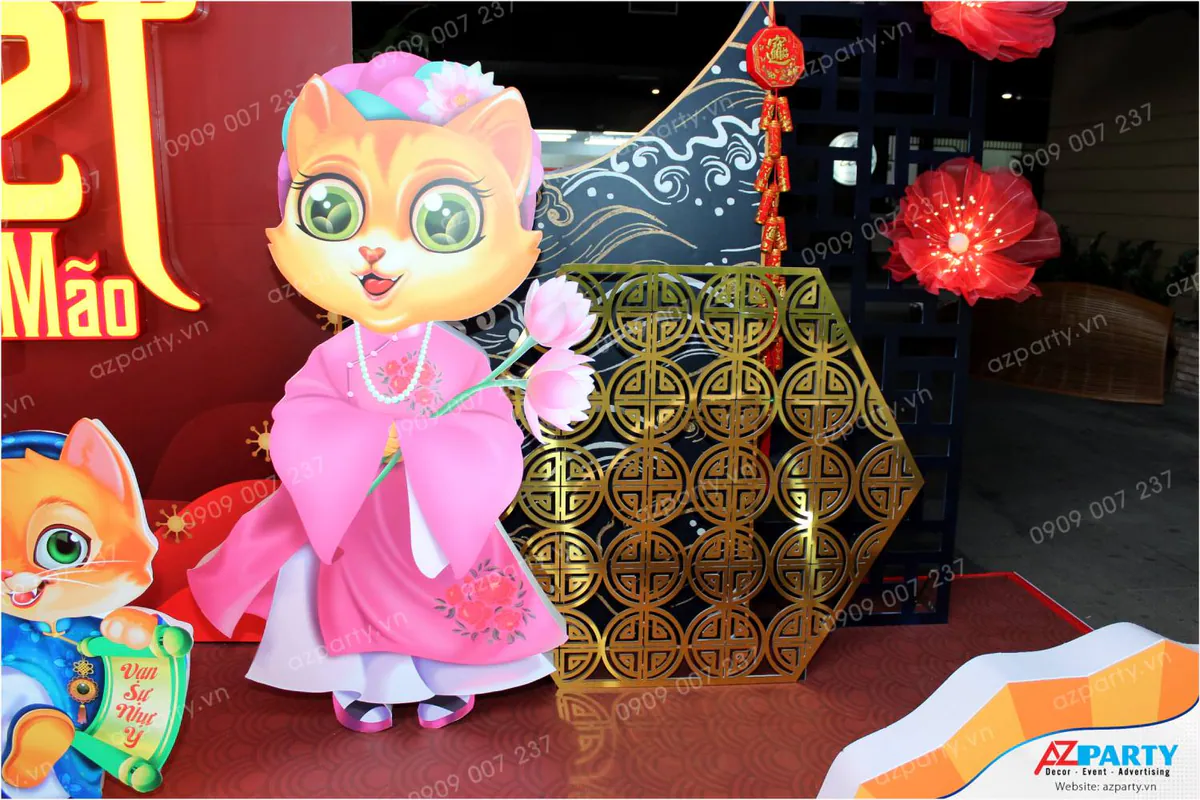Mô hình mèo mẹ duyên dáng mặc áo dài hồng cầm hoa sen trang trí cạnh tấm vách hoa văn đồng tiền mạ vàng lấp lánh