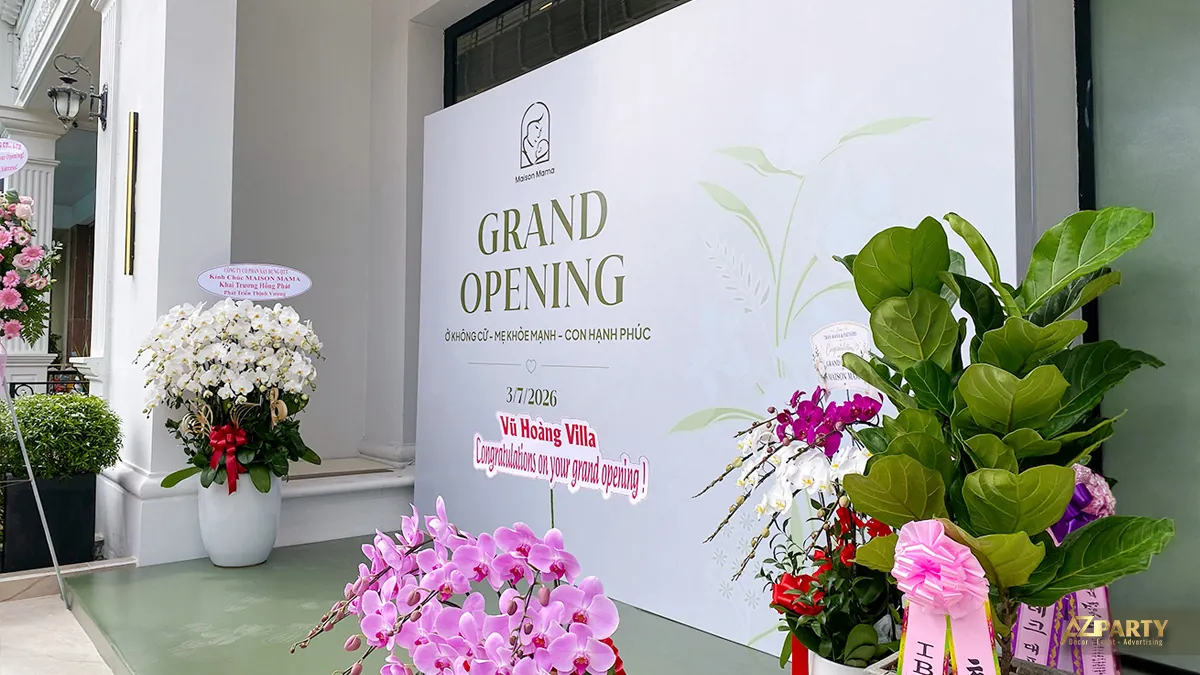 Góc nghiêng backdrop khai trương Maison Mama với sàn xanh, vách Grand Opening và các lẵng hoa chúc mừng