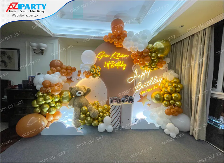 Trang trí tiệc sinh nhật kết hợp cho bé Gia Khiêm 1 tuổi và Gia Minh 5 tuổi với backdrop 3D hình gấu teddy khổng lồ, thảm cỏ xanh, các chữ neon LED rực rỡ và viền bóng bay nghệ thuật tông màu cam, trắng, nâu ấm áp.
