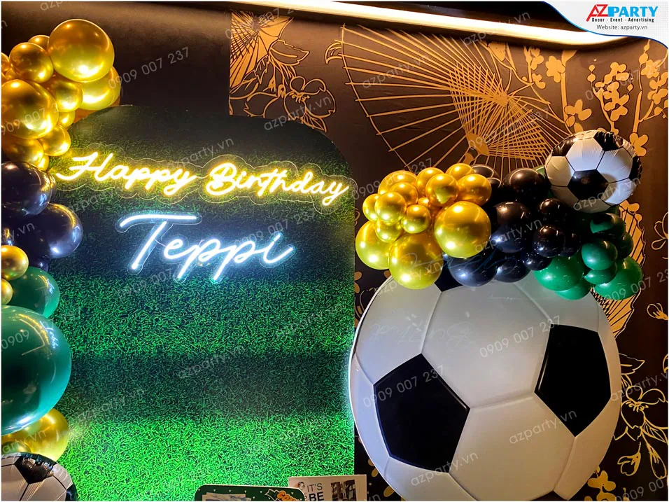 Cận cảnh chữ đèn neon Happy Birthday Teppi phát sáng rực rỡ trên nền backdrop mô phỏng thảm cỏ sân bóng màu xanh lá.