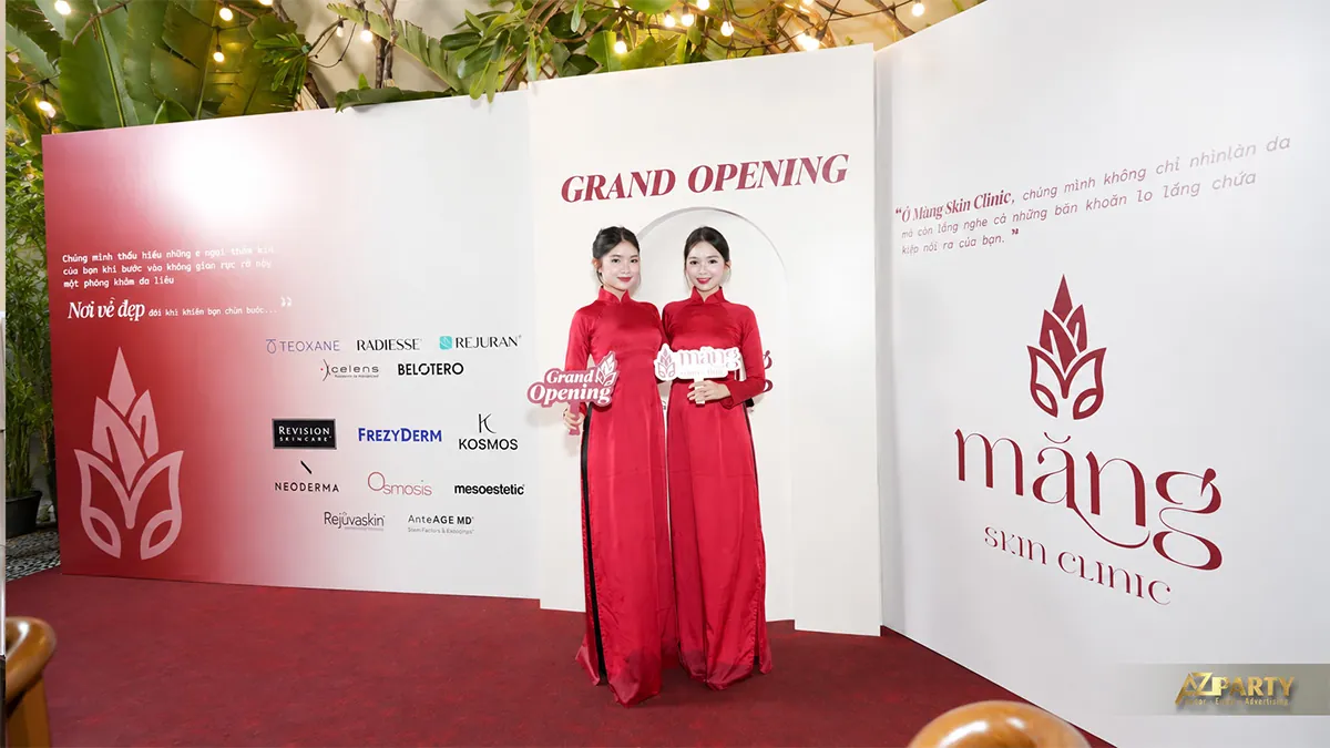 Hai người mặc áo dài đỏ cầm bảng check-in trước backdrop