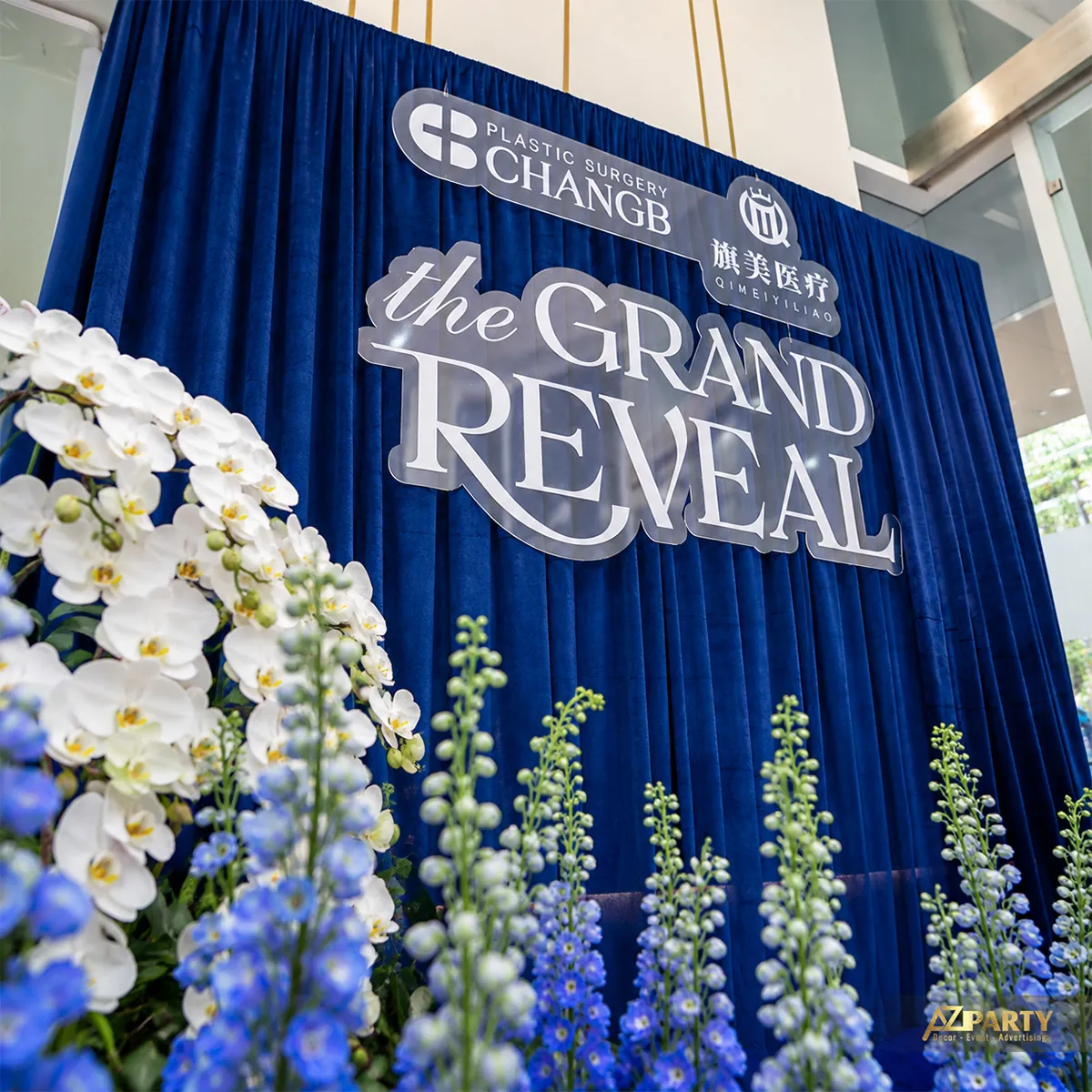 Cận chữ The Grand Reveal giữa hoa lan trắng và hoa xanh