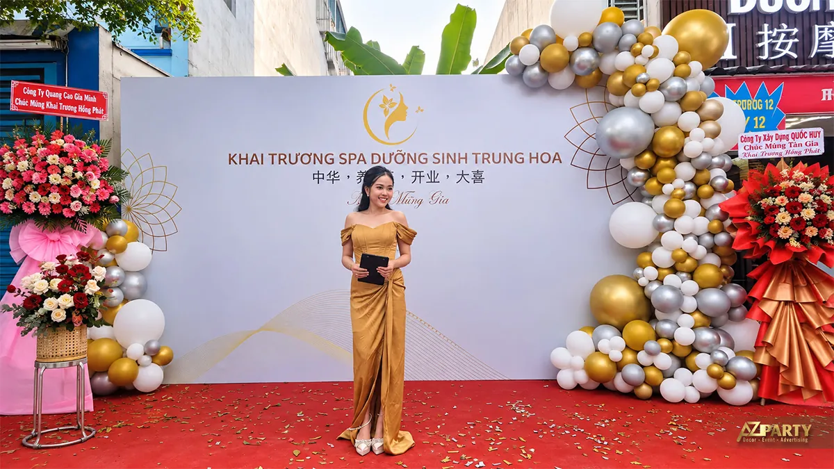 Người dẫn chương trình mặc váy vàng trước backdrop spa