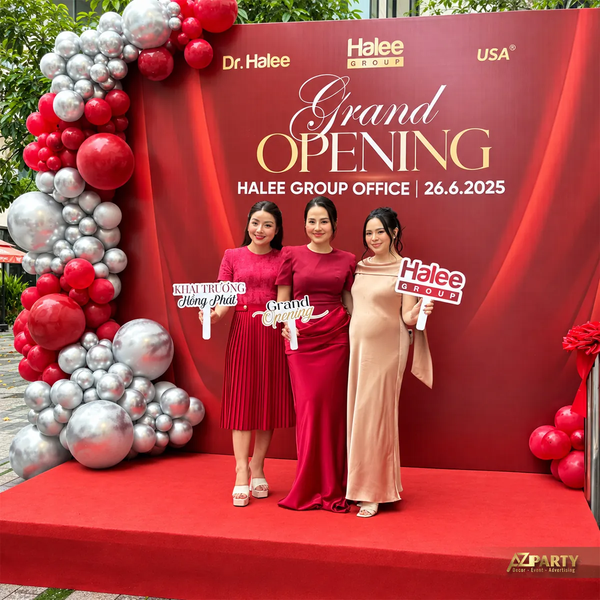 Khách mời cầm bảng chúc mừng trước backdrop khai trương
