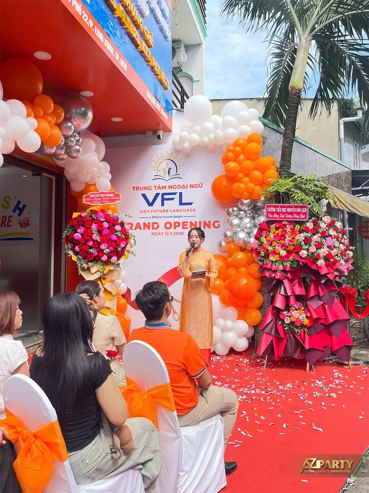 Người dẫn chương trình mặc áo dài vàng phát biểu trước backdrop khai trương