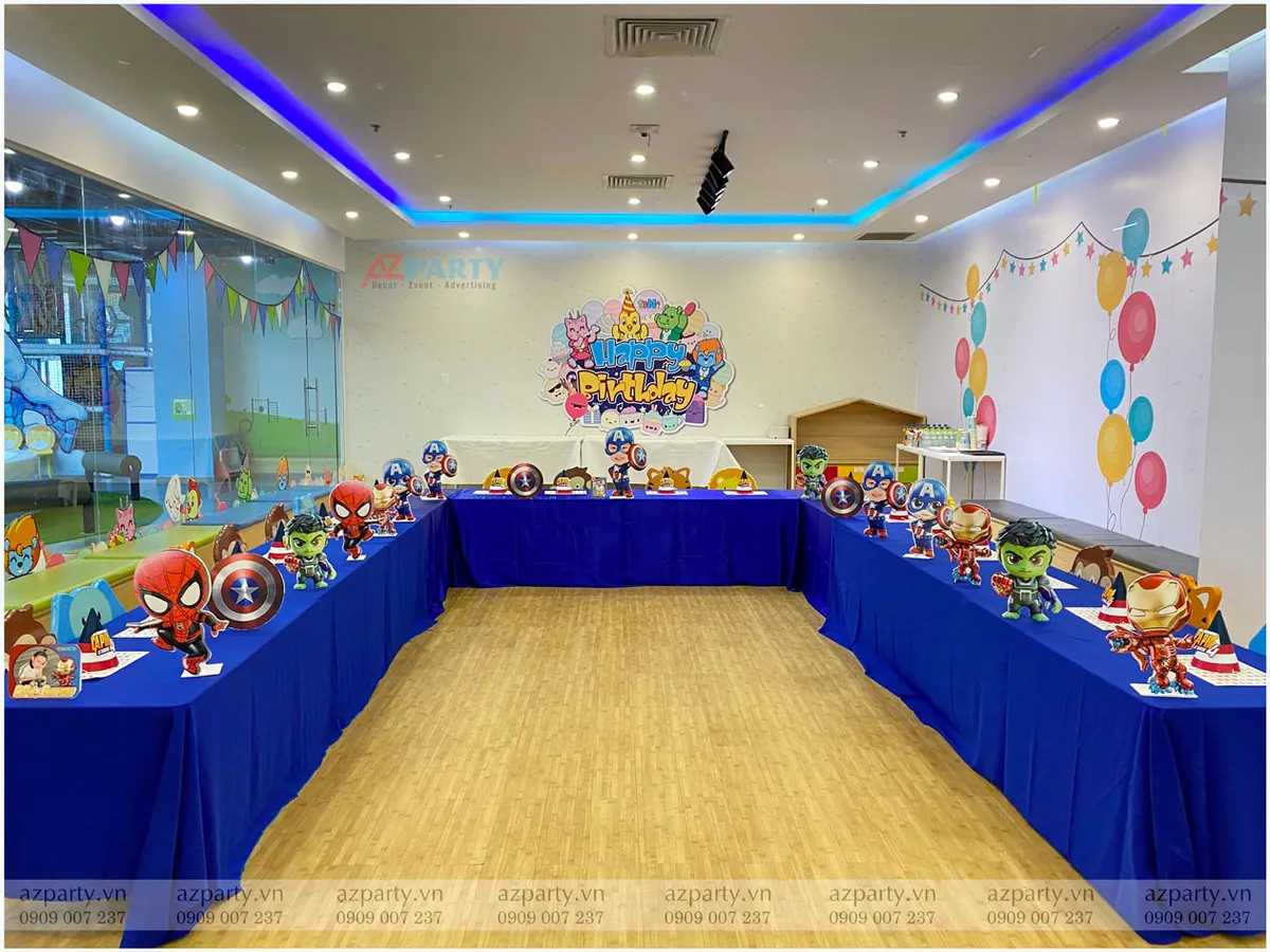 Bàn tiệc sinh nhật cho bé được phủ khăn xanh dương nổi bật, dọc theo mép bàn trang trí các mô hình mini Avengers như Iron Man, Thor, Captain America.