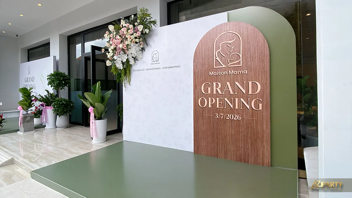 Góc rộng không gian khai trương Maison Mama với backdrop xanh sage, bảng Grand Opening và cụm hoa tươi bên trái