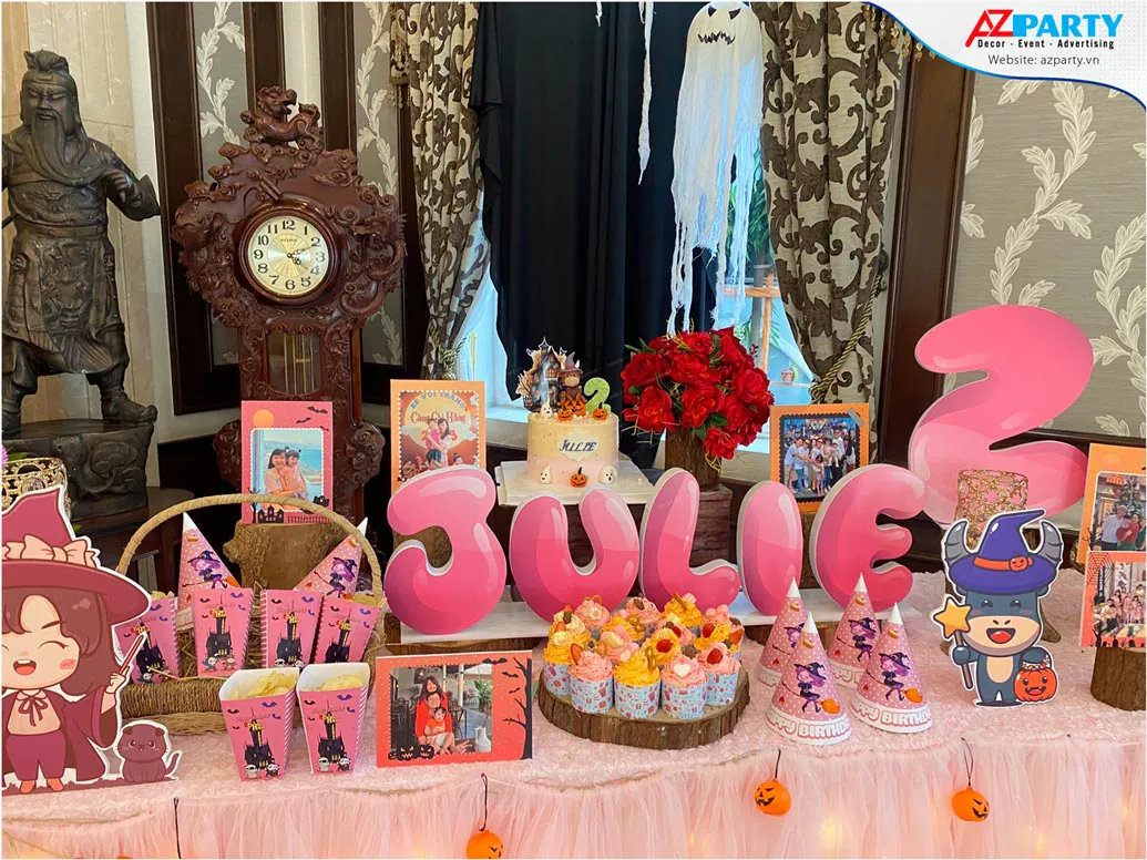 Cận cảnh bàn tiệc ngọt của bé Julie nổi bật với chữ xốp tên bé màu hồng 3D đặt cạnh đĩa bánh cupcake và nón phù thủy bằng giấy.