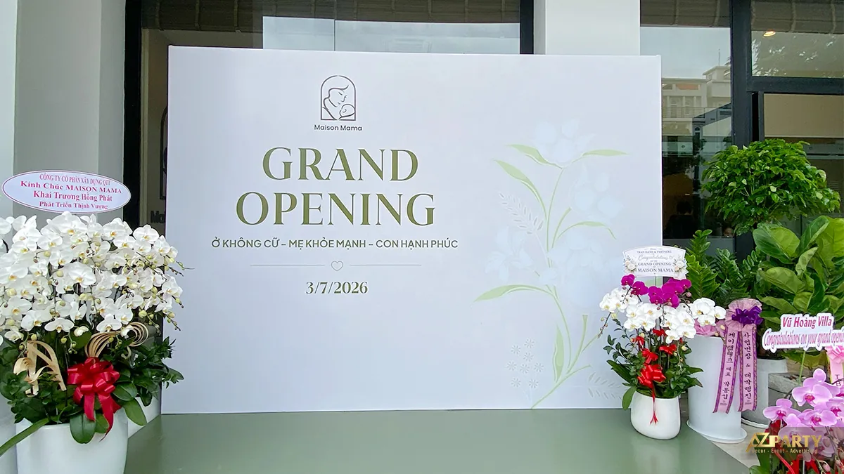 Khu check-in Maison Mama với backdrop Grand Opening nền trắng xanh, hoa chúc mừng và sàn xanh