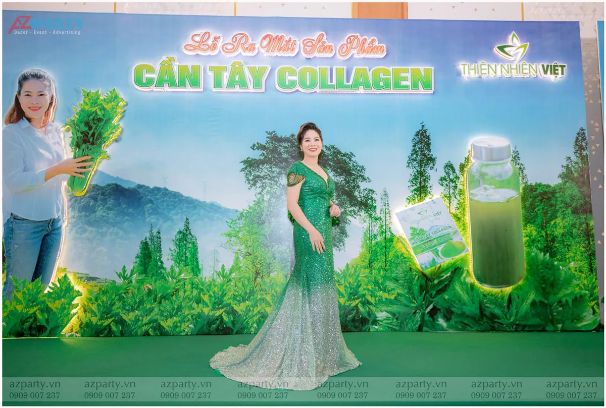 Nữ đại sứ hoặc khách mời trong trang phục đầm dạ hội xanh lấp lánh tạo dáng trước backdrop lễ ra mắt Cần Tây Collagen.