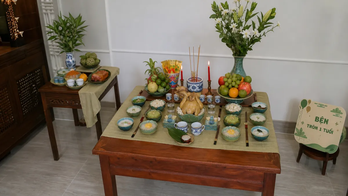 Mâm thôi nôi trên bàn gỗ gồm gà luộc, các chén xôi chè, hai mâm trái cây và hoa trắng