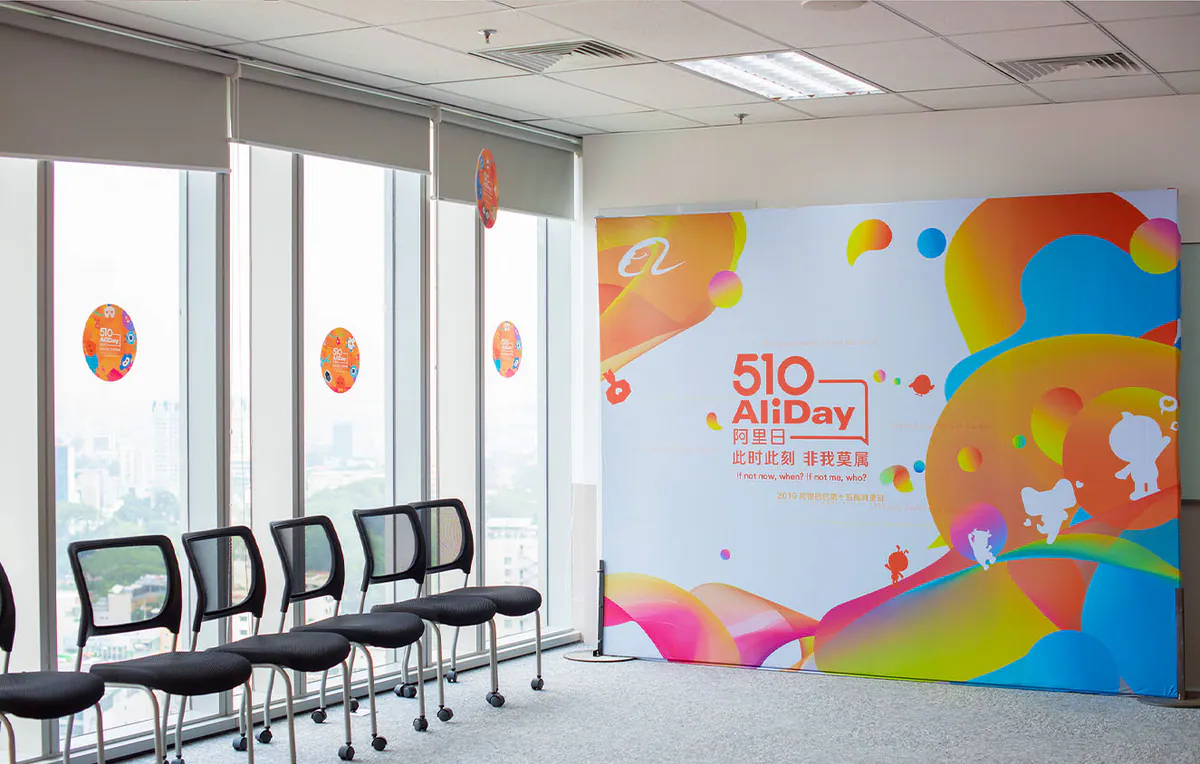 Khu vực sân khấu trong văn phòng với backdrop sự kiện 510 AliDay thiết kế rực rỡ và hàng ghế chờ bên cửa kính.