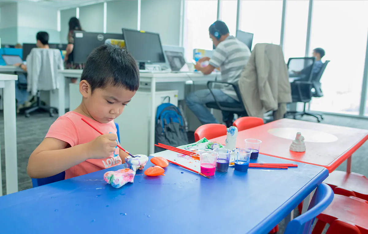 Một bé trai ngồi tại bàn nhựa màu xanh dương đang say sưa dùng cọ vẽ tô màu cho tượng thạch cao, phía sau là không gian làm việc của các nhân viên văn phòng Aliday.