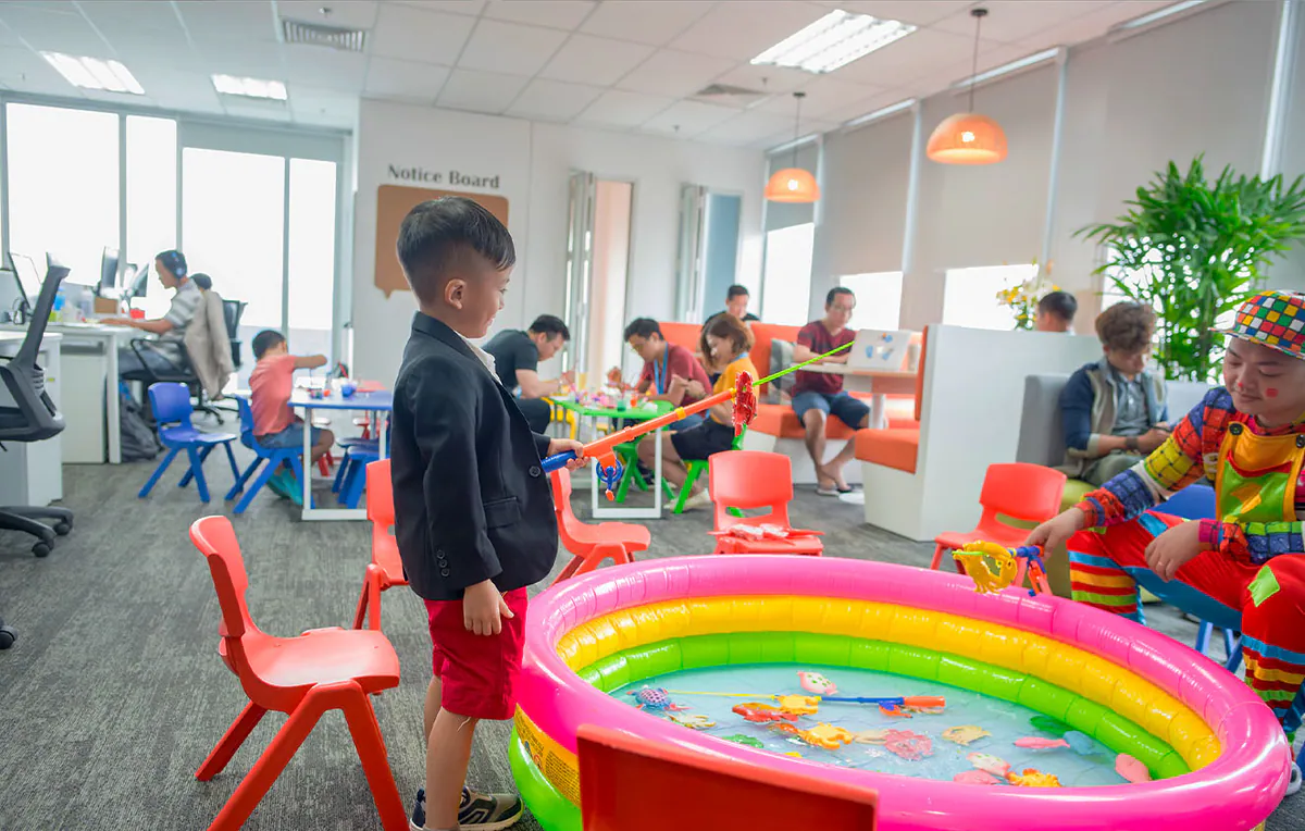 Bé trai mặc vest đen lịch lãm đang chơi câu cá nhựa tại bể phao nhiều màu sắc cùng chú hề trang phục rực rỡ tại ngày hội gia đình Aliday.
