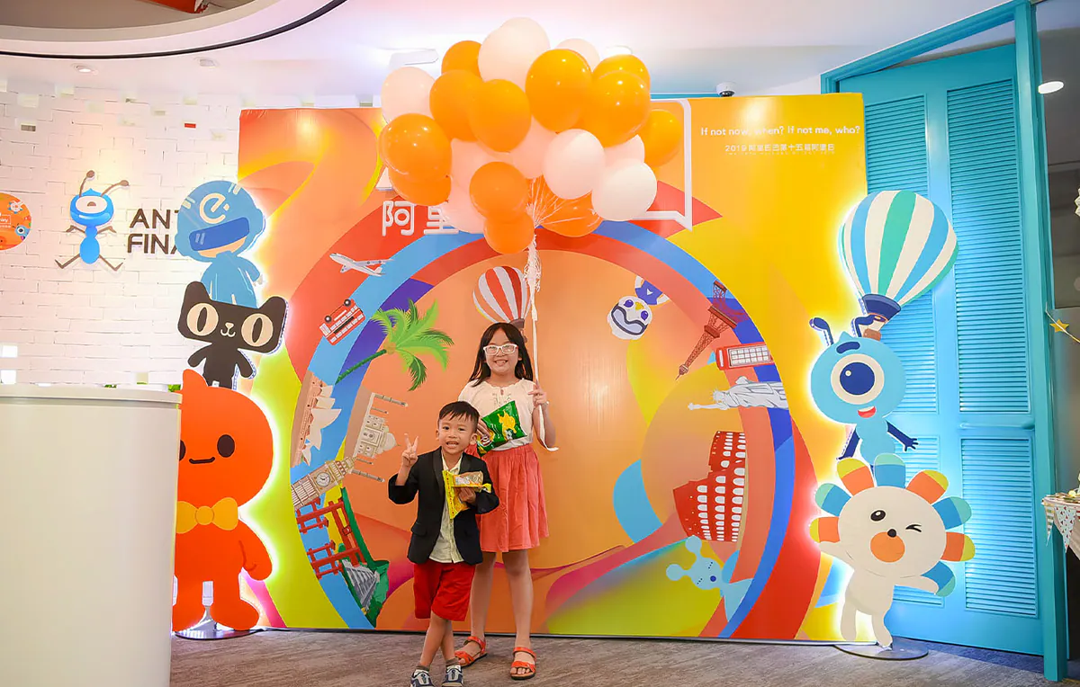 Hai bé thiếu nhi đứng chụp hình kỷ niệm vui tươi trước backdrop sự kiện 510 AliDay rực rỡ có trang trí bóng bay màu vàng cam và trắng.