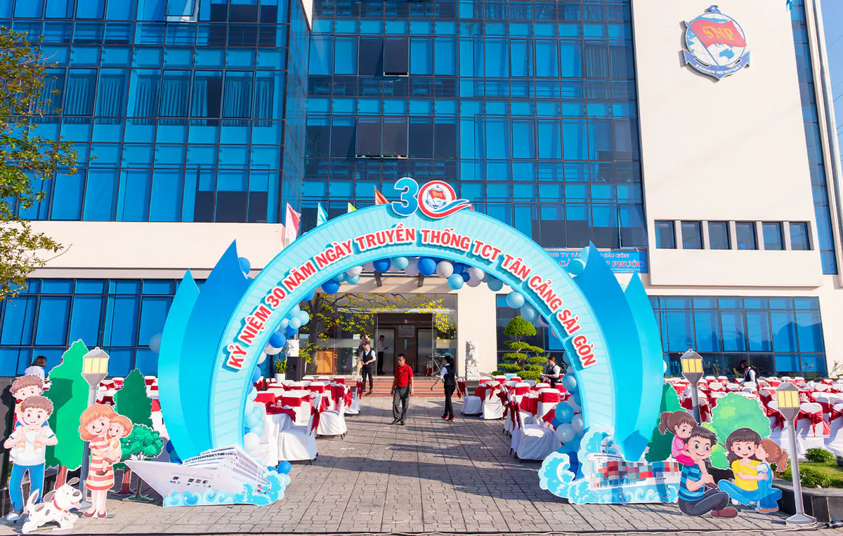 Cổng chào sự kiện Kỷ niệm 30 năm Ngày truyền thống TCT Tân Cảng Sài Gòn vào ban ngày, xung quanh bố trí nhiều bàn ghế tiệc phủ khăn trắng đỏ chu đáo.