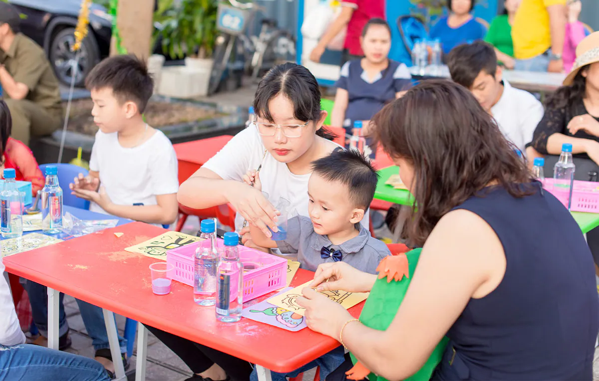 Người mẹ trẻ ân cần giúp con trai nhỏ chọn màu cát để đổ lên bức tranh khỉ dễ thương tại ngày hội.