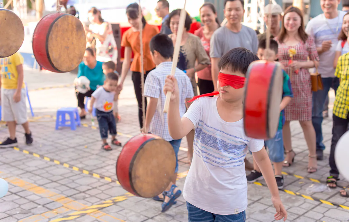 Một bé trai bịt mắt bằng vải đỏ đang tập trung dùng gậy để tham gia trò chơi bịt mắt đập trống dân gian náo nhiệt tại Ngày hội gia đình.