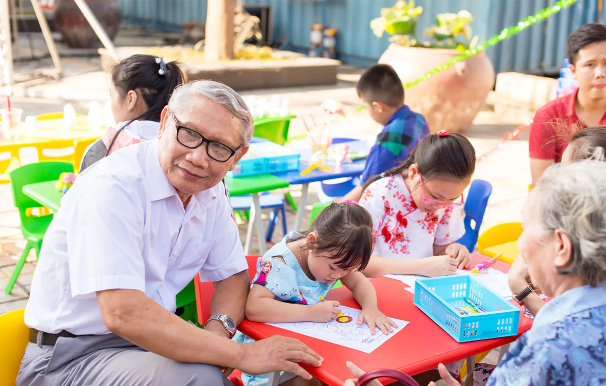 Ông nội hoặc ông ngoại lớn tuổi đeo kính, cười hiền hậu khi nhìn cháu gái đang say sưa tô màu tại sự kiện Ngày hội gia đình.