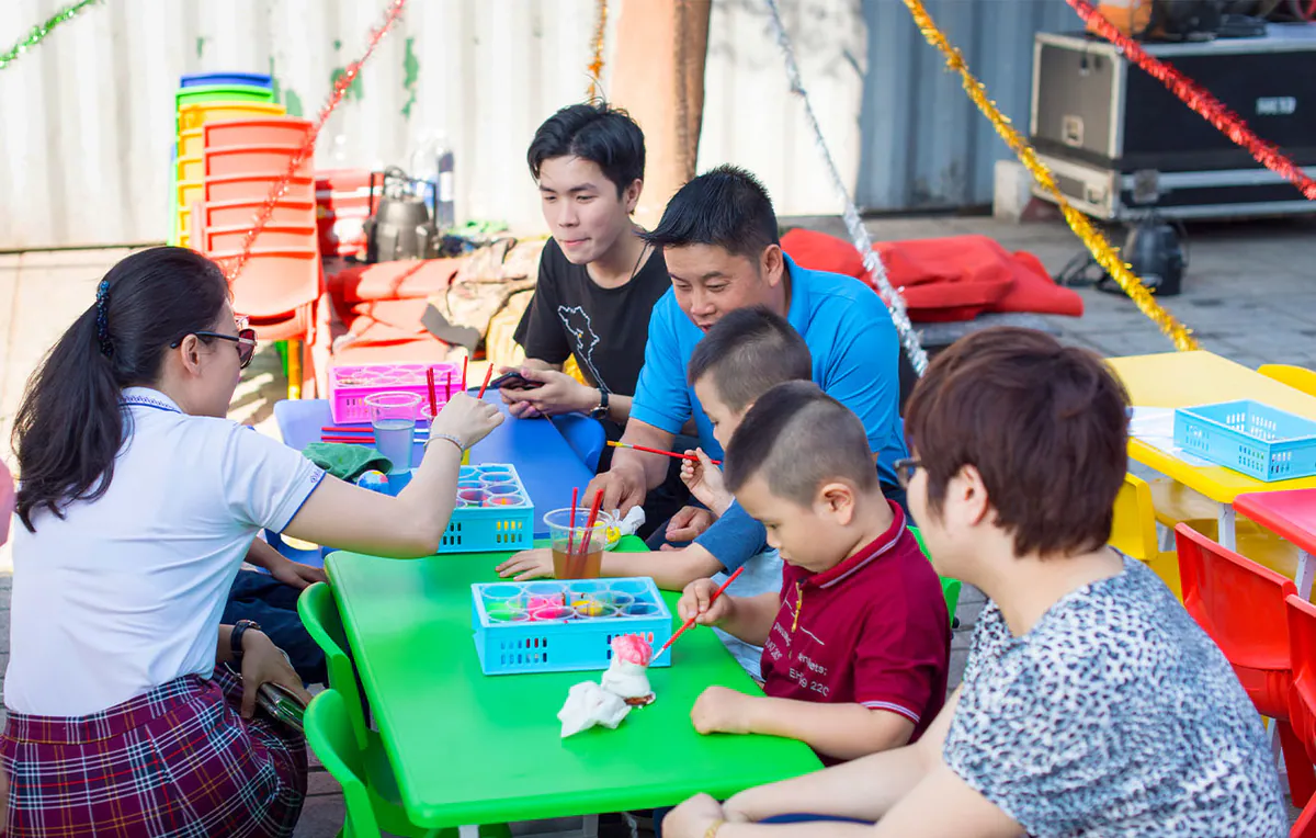 Các em nhỏ cùng ba mẹ tập trung xung quanh bàn nhựa nhỏ màu xanh lá để cùng nhau tô tượng thạch cao vui vẻ trong không khí ngày hội.