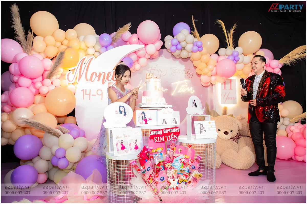 Bé Kim Tiền mặc váy tím lấp lánh đội vương miện nhỏ cười rạng rỡ cắt bánh sinh nhật cùng nam MC hoạt náo trên sân khấu.