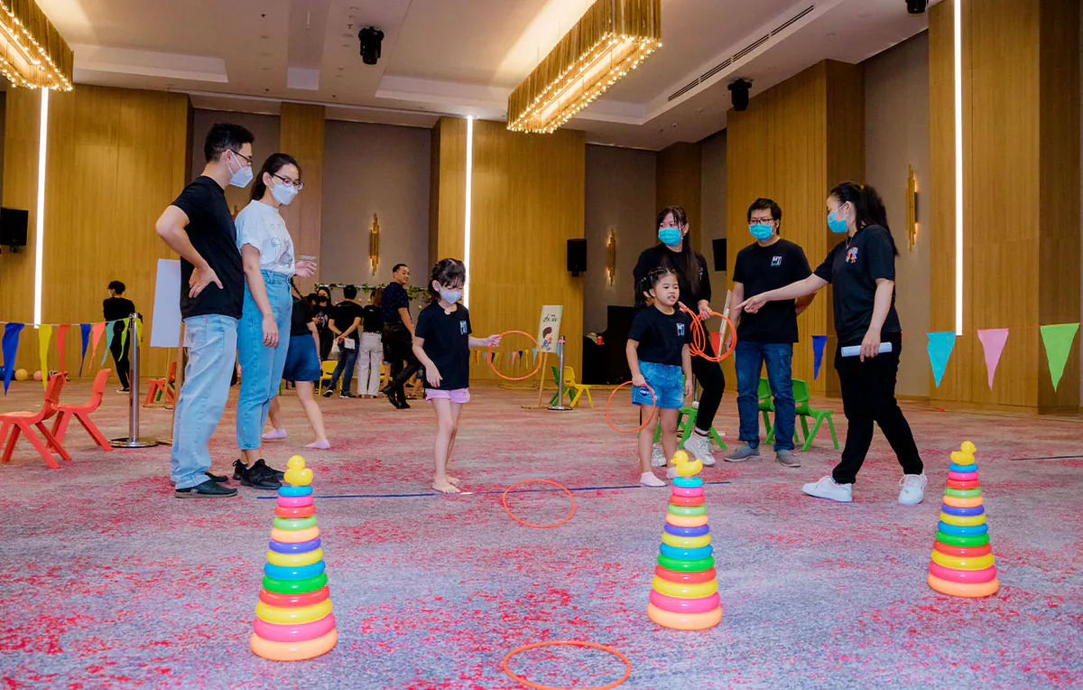 Các bé gái đeo khẩu trang đang hào hứng ném các vòng nhựa màu cam vào cọc ném hình tháp nhiều màu sắc tại sảnh tiệc của Holiday Inn.