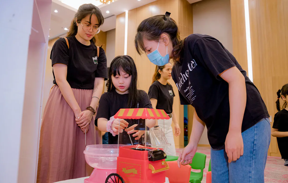 Một bé gái đang tự tay làm kẹo bông gòn ngọt ngào bằng máy làm kẹo bông mini dưới sự hướng dẫn tỉ mỉ của các nữ nhân viên.