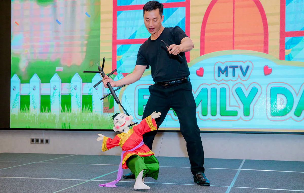 Nghệ sĩ múa rối mặc đồ đen đang biểu diễn điều khiển con rối chú Tễu mặc áo đỏ quần xanh lá trên sân khấu chính của sự kiện.