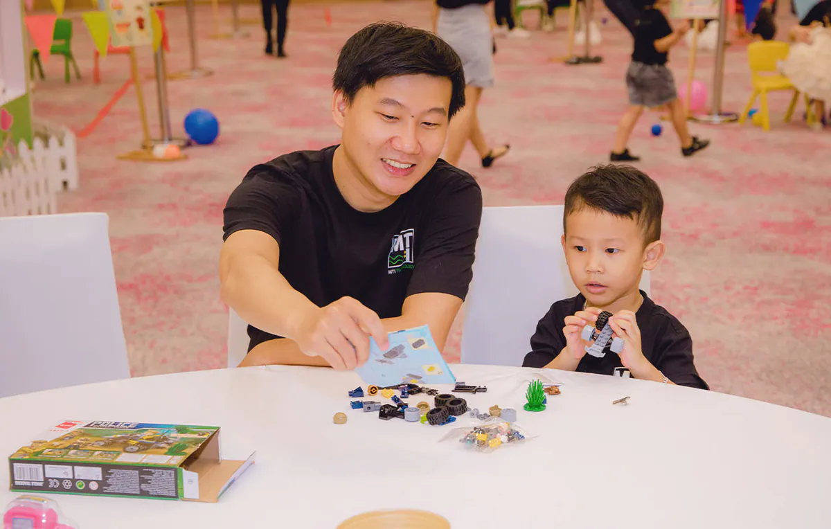 Người cha cười tươi khi chỉ cho con trai nhỏ cách lắp ráp đồ chơi Lego mô hình xe cảnh sát tại bàn hoạt động của ngày hội gia đình.