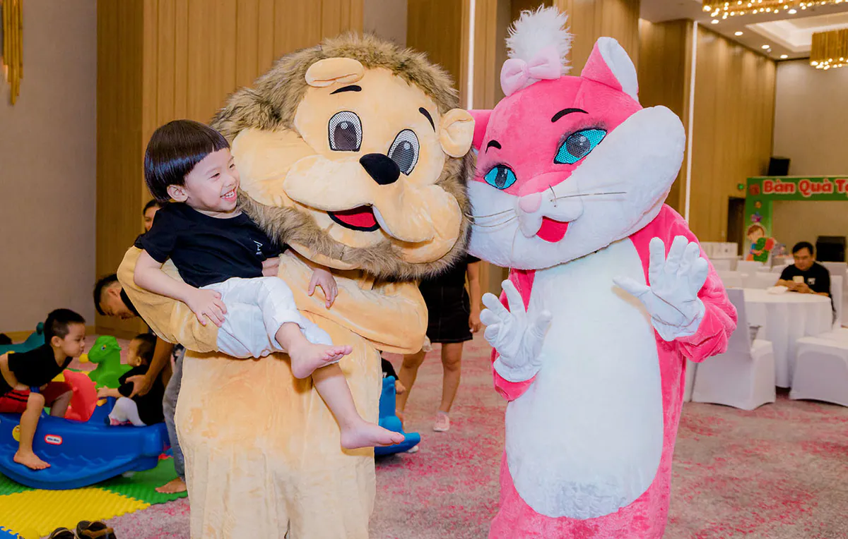 Mascot chú sư tử vàng dễ thương đang bế một bé trai cười thích thú bên cạnh mascot mèo hồng xinh xắn đang vẫy tay chào tại ngày hội.