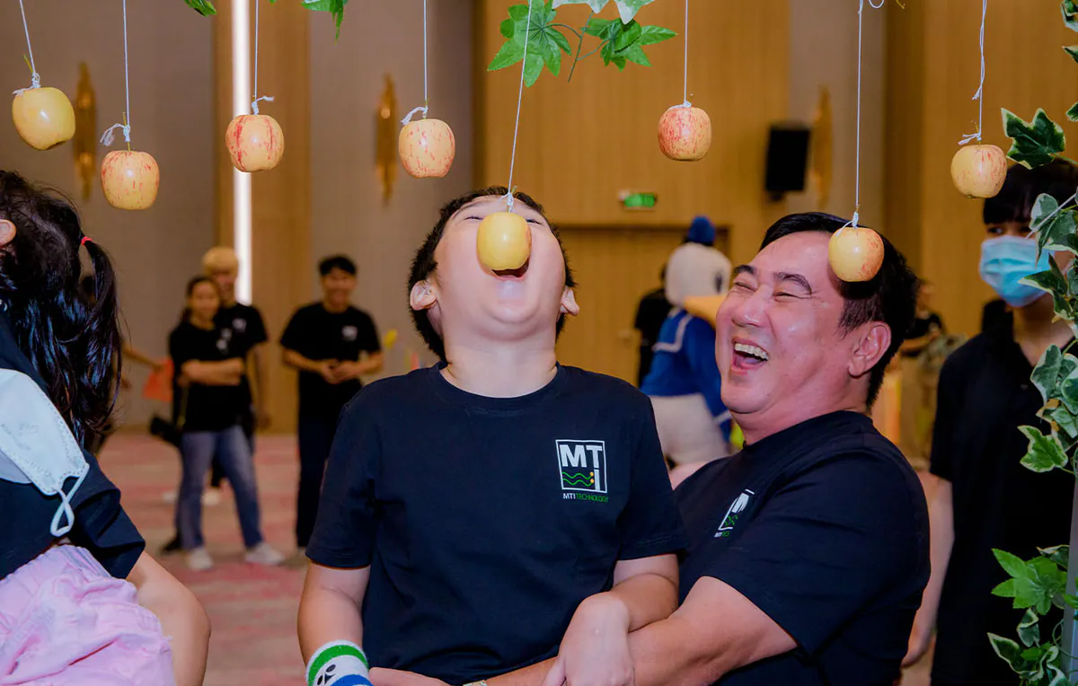 Một người bố đang nâng con trai mình lên để bé tham gia trò chơi cắn táo treo dây đầy kịch tính và vui nhộn.