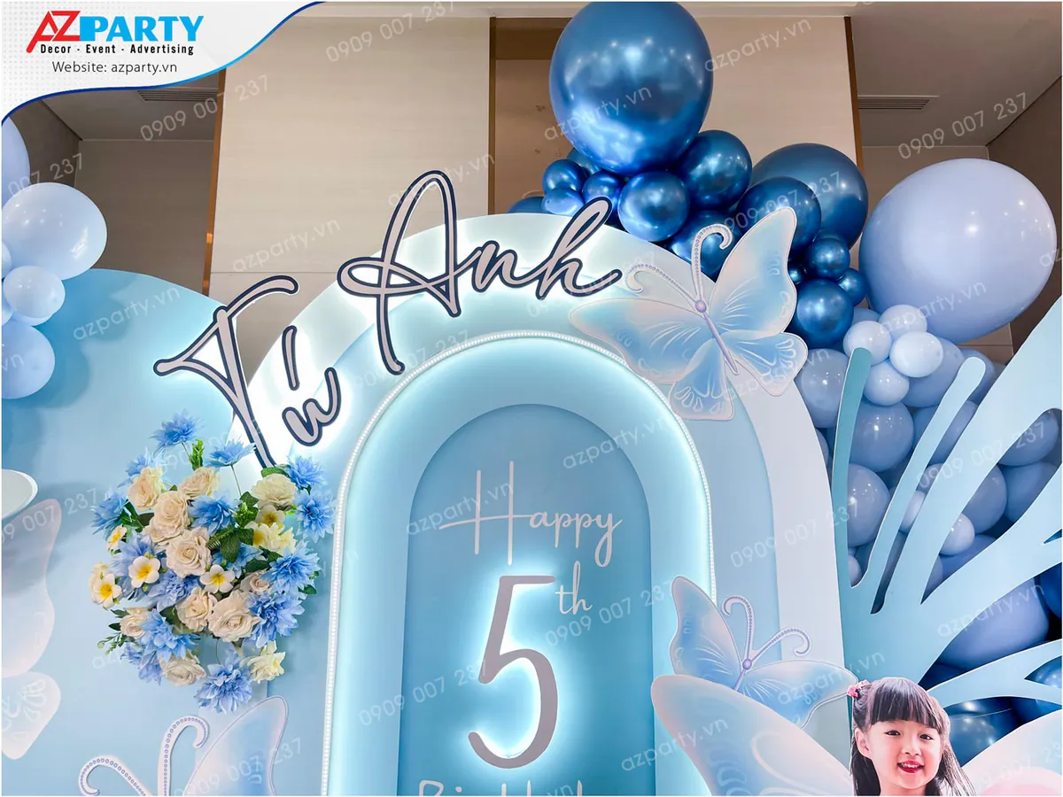 Cận cảnh bảng tên hiệu ứng đèn LED sáng 'Tú Anh' và dòng chữ 'Happy 5th' trên nền backdrop hình cổng vòm màu xanh dương tinh tế kết hợp hoa tươi cùng tông.