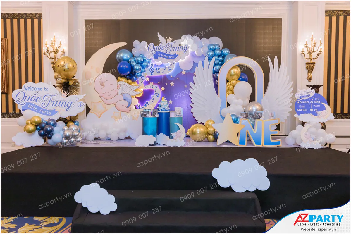 Trang trí sân khấu thôi nôi bé Quốc Trung chủ đề thiên thần nhỏ ngủ trên mặt trăng lãng mạn với đôi cánh thiên thần lớn và bóng bay lấp lánh.