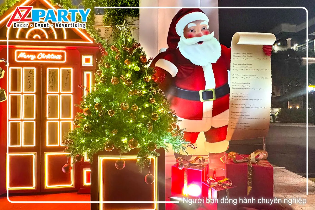 cận cảnh cây thông noel với quả châu đỏ cùng dây đèn vàng, bên cạnh là ông già noel vác quà, tay còn lại cầm danh sách các trẻ em được nhận quà tặng