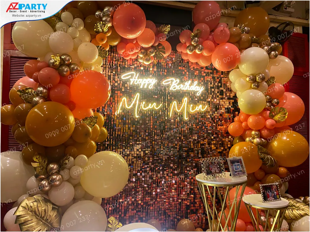 Cận cảnh chữ đèn neon Happy Birthday Miu Miu nổi bật trên nền backdrop sequin phản chiếu ánh sáng rực rỡ và cụm bong bóng nghệ thuật nhiều màu sắc.