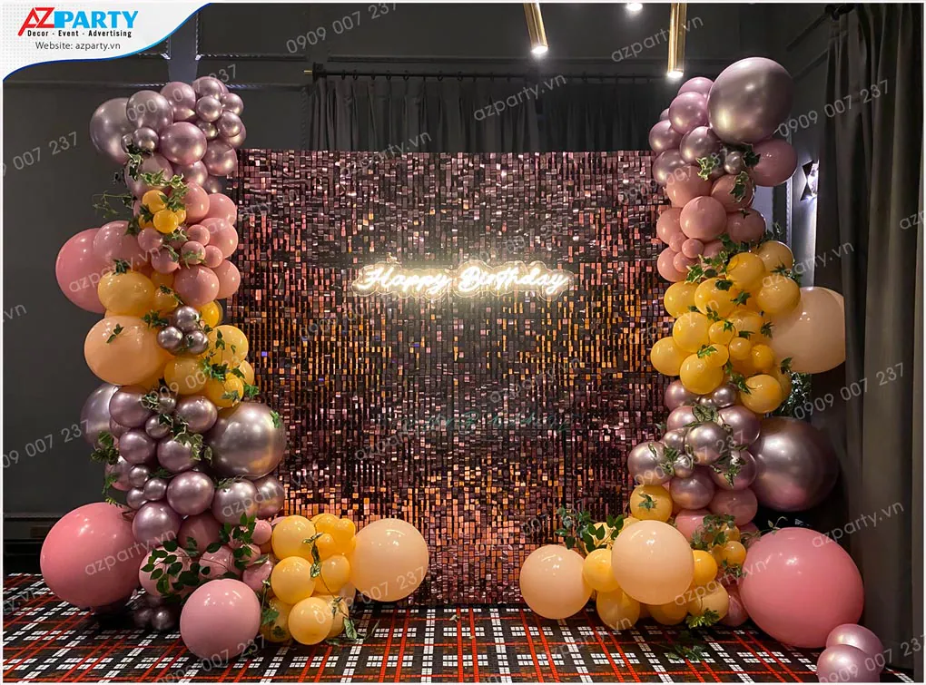 Sân khấu trang trí tiệc sinh nhật lung linh với backdrop sequin đồng, chữ neon Happy Birthday và hai dải bong bóng nghệ thuật màu tím metallic, hồng và vàng nhạt.