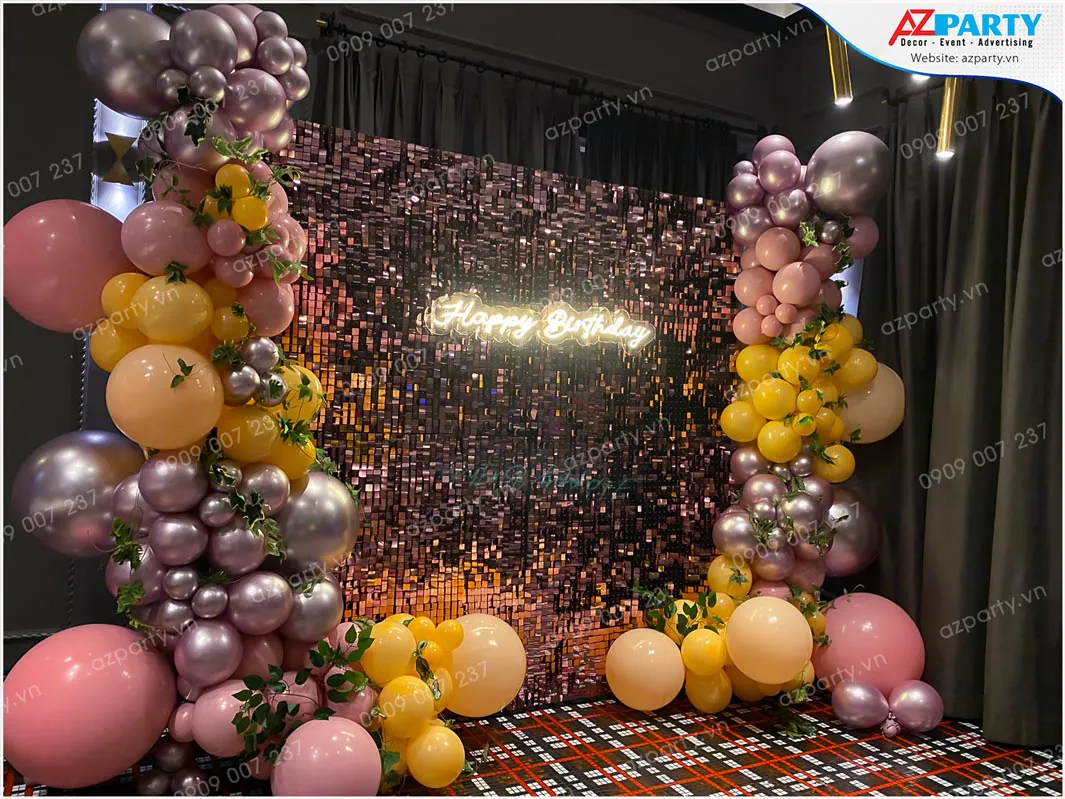Góc chụp chính diện backdrop sequin lấp lánh phối cùng bong bóng màu pastel, bóng chrome và nhánh lá xanh trang trí tự nhiên.